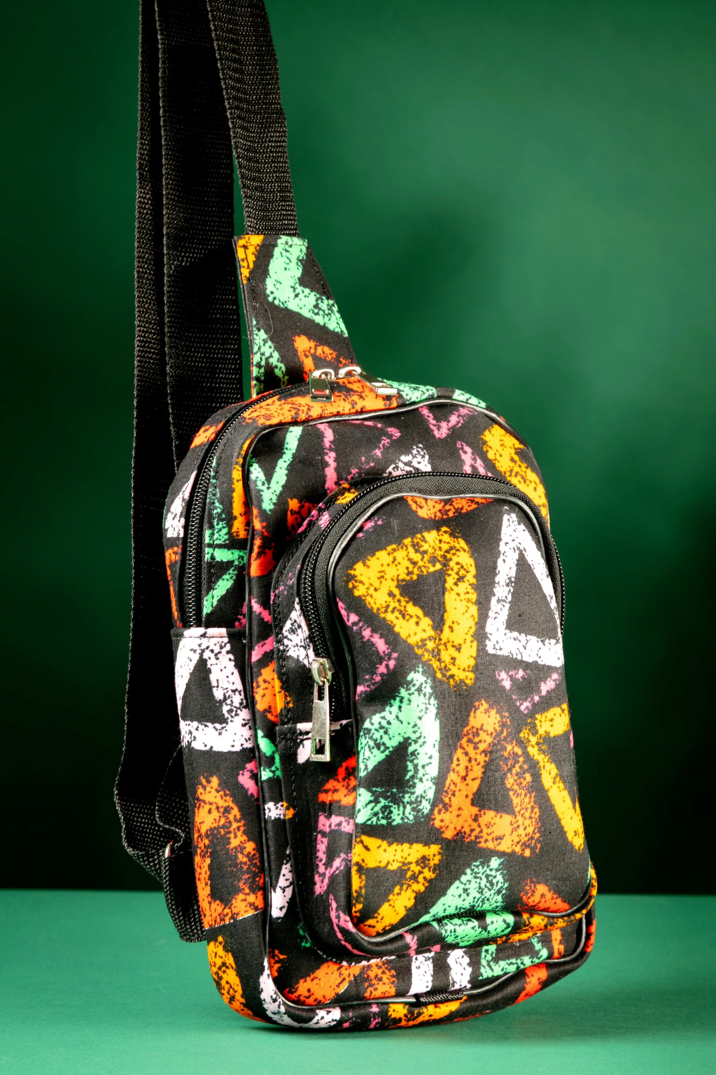 Angled view of the Adungu African Print Chest Fanny with bold multicolored triangular patterns, black adjustable strap, and front zipper pocket, displayed against a green background.