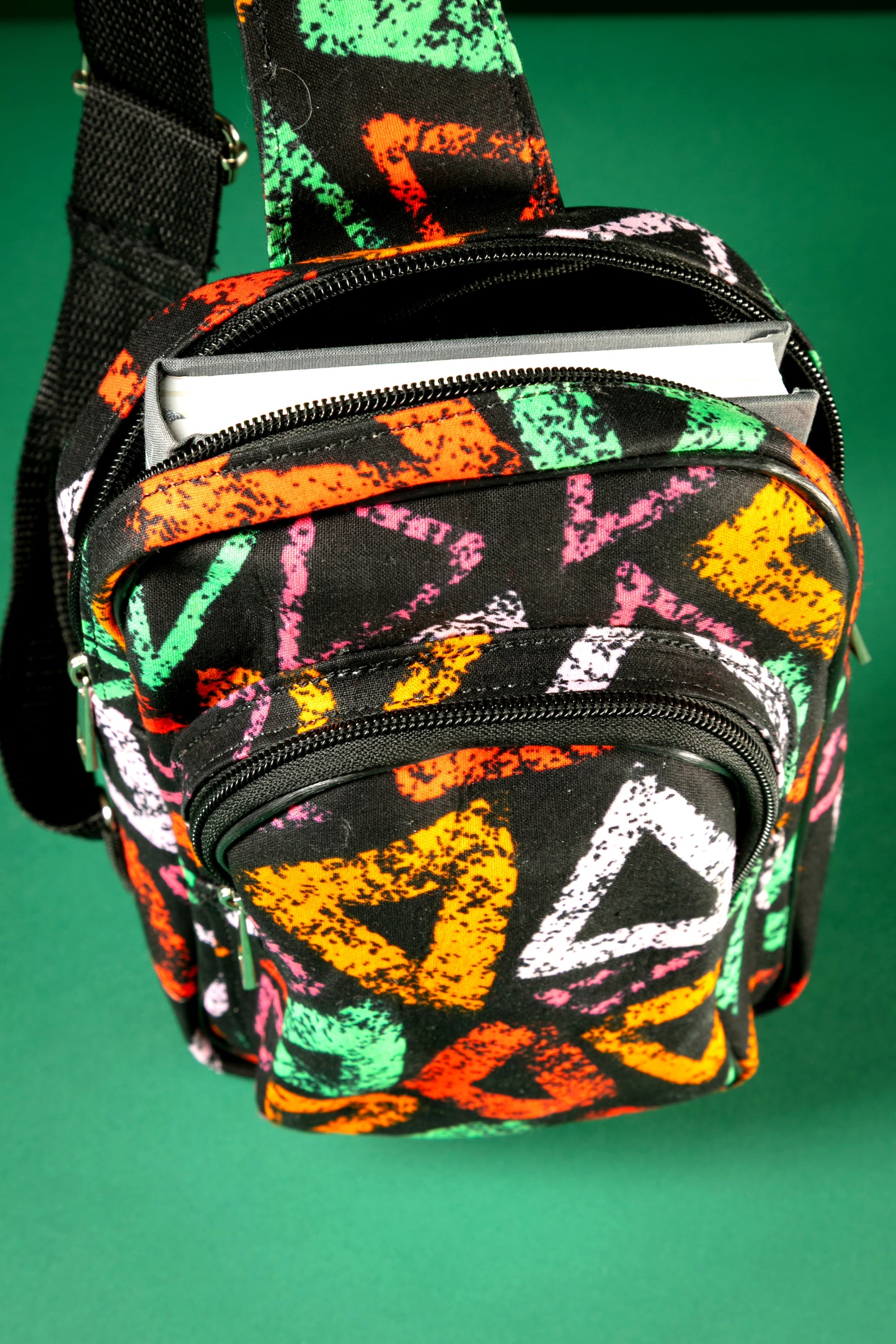 Top view of the Adungu African Print Chest Fanny with bold multicolored triangular patterns, black adjustable strap, and front zipper pocket, displayed against a green background.