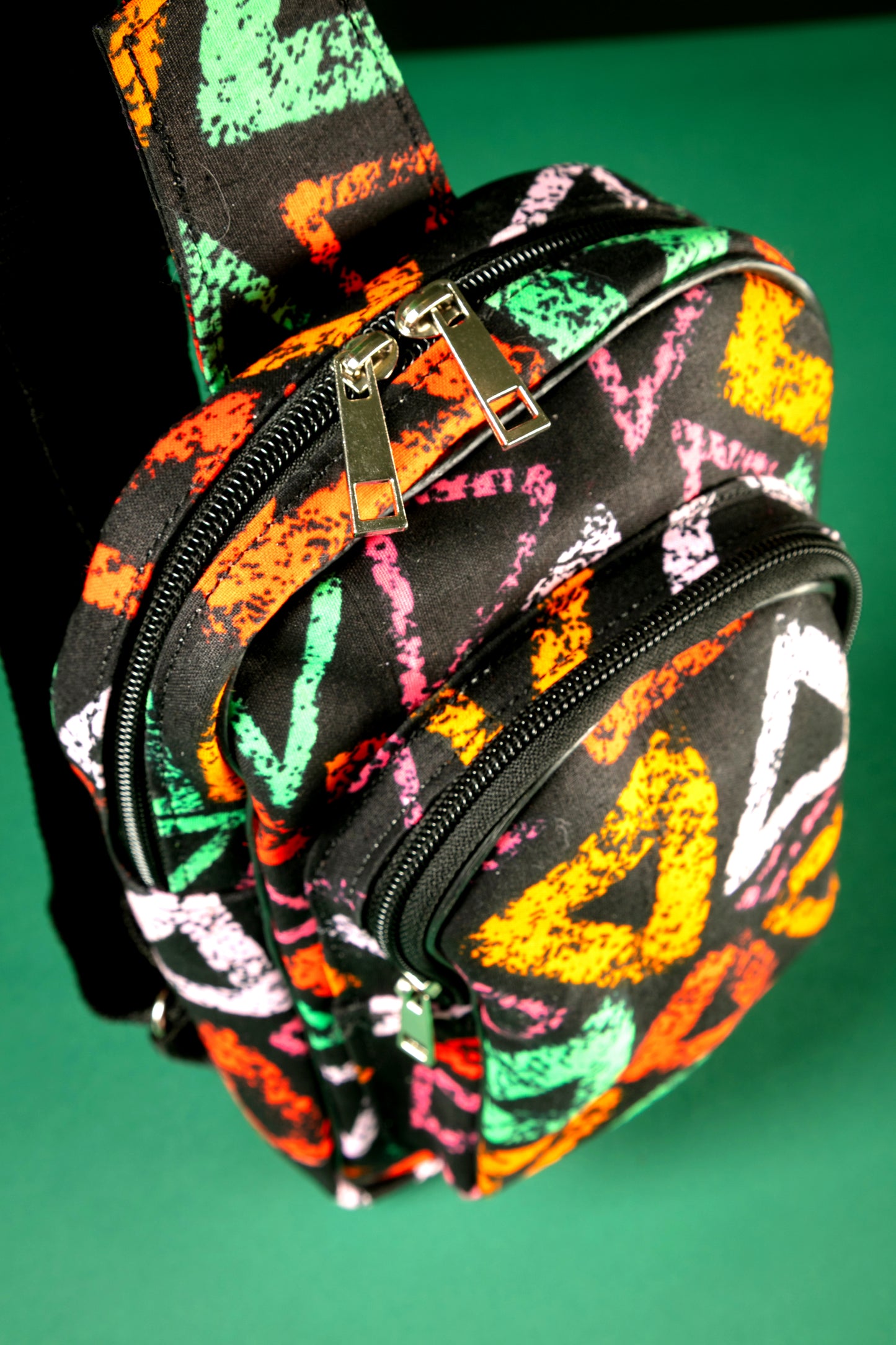 Top close-up view of the Adungu African Print Chest Fanny with bold multicolored triangular patterns, black adjustable strap, and front zipper pocket, displayed against a green background.