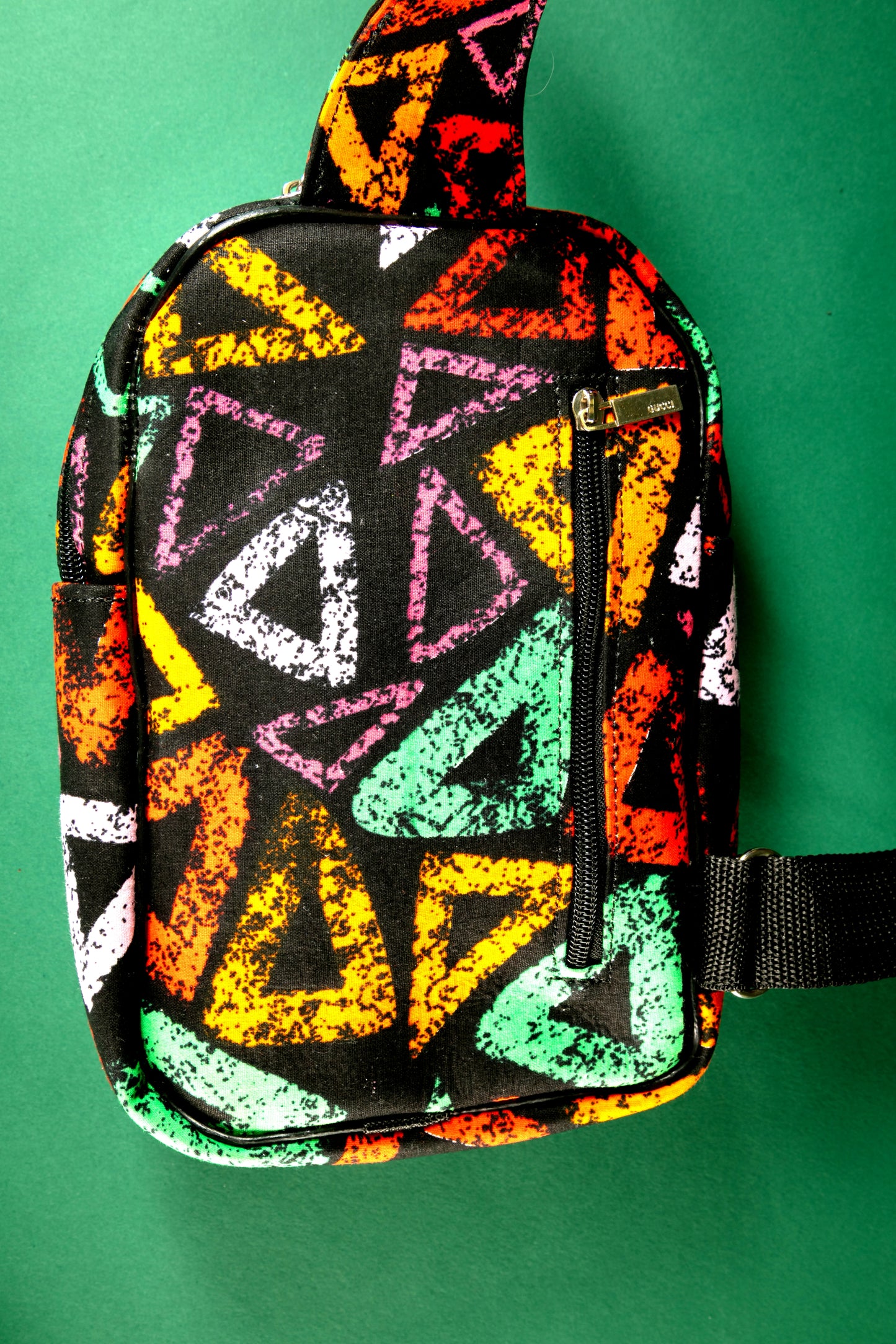 Back view of the Adungu African Print Chest Fanny with bold multicolored triangular patterns, black adjustable strap, and front zipper pocket, displayed against a green background.