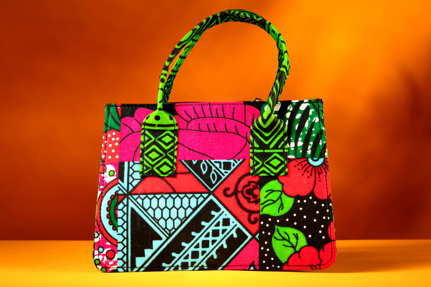 Front view of the Balangi Bag – a structured African print handbag featuring a multicolor design with contrasting green patterned handles, photographed against a warm orange background.