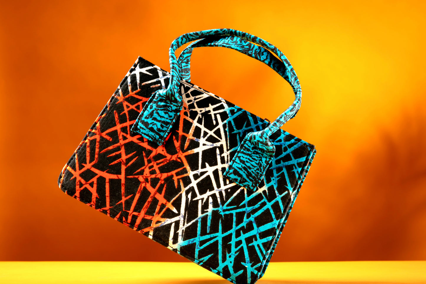Front view of the Gyil Bag – a structured African print handbag featuring a multicolor design with contrasting blue patterned handles, photographed against a warm orange background.