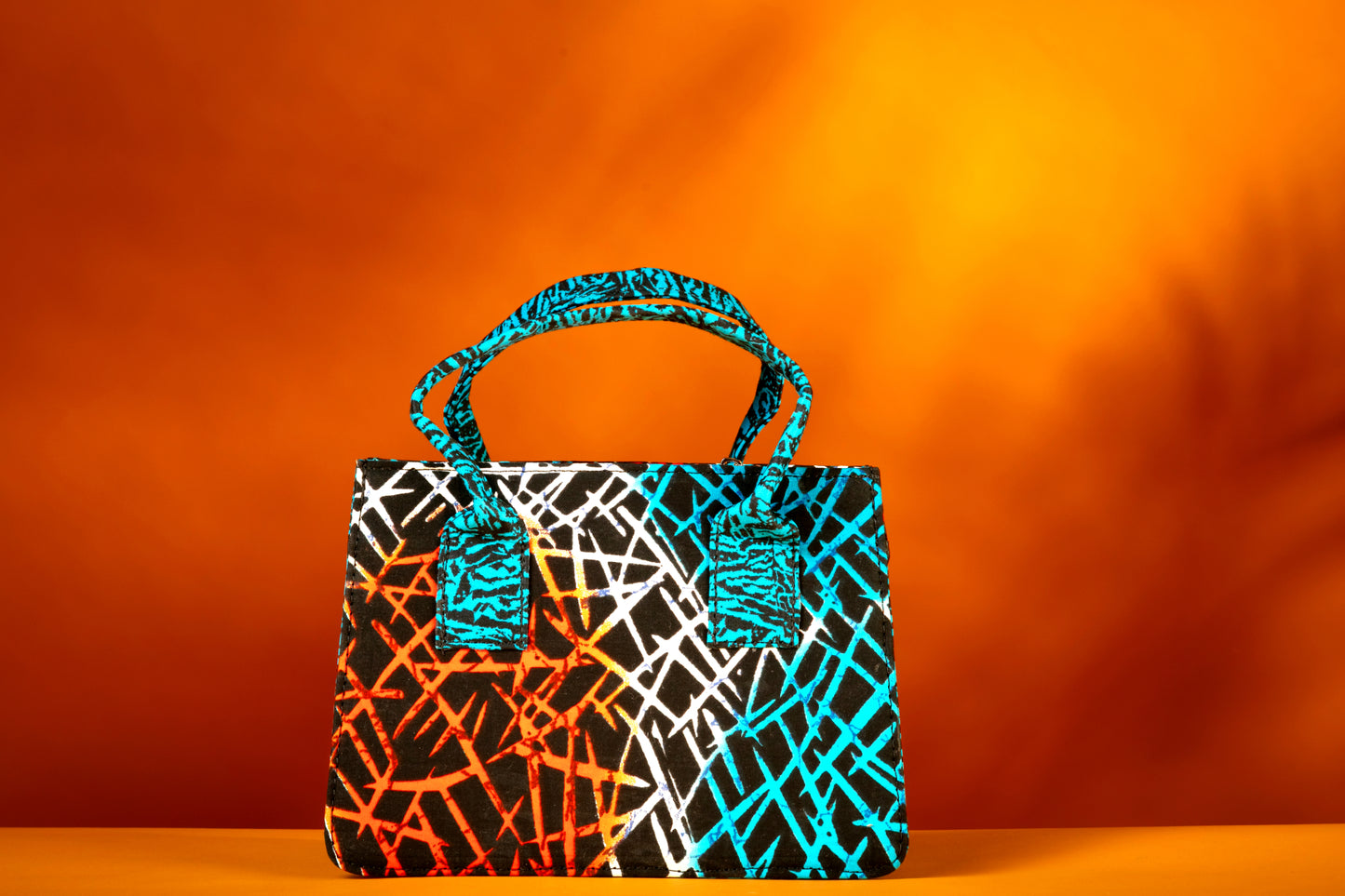 Front view of the Gyil Bag – a structured African print handbag featuring a multicolor design with contrasting blue patterned handles, photographed against a warm orange background.