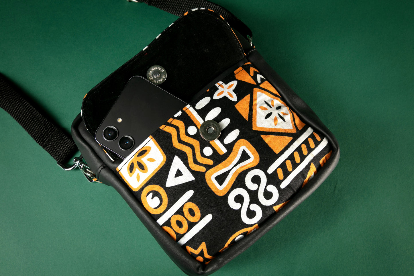 Flat lay of a black crossbody bag with brown, black & white tribal patterns with the flap to the front pocket opened on a green background