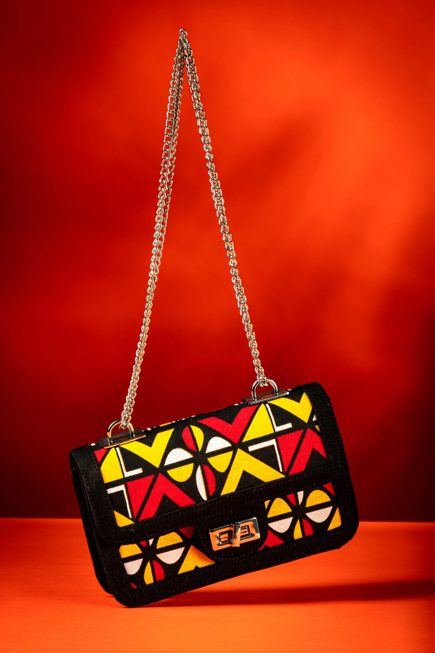 Shekere African print handbag with yellow, red, black and white Ankara fabric and black denim trim, featuring a silver chain strap and chrome twist-lock closure, handmade in Nigeria.