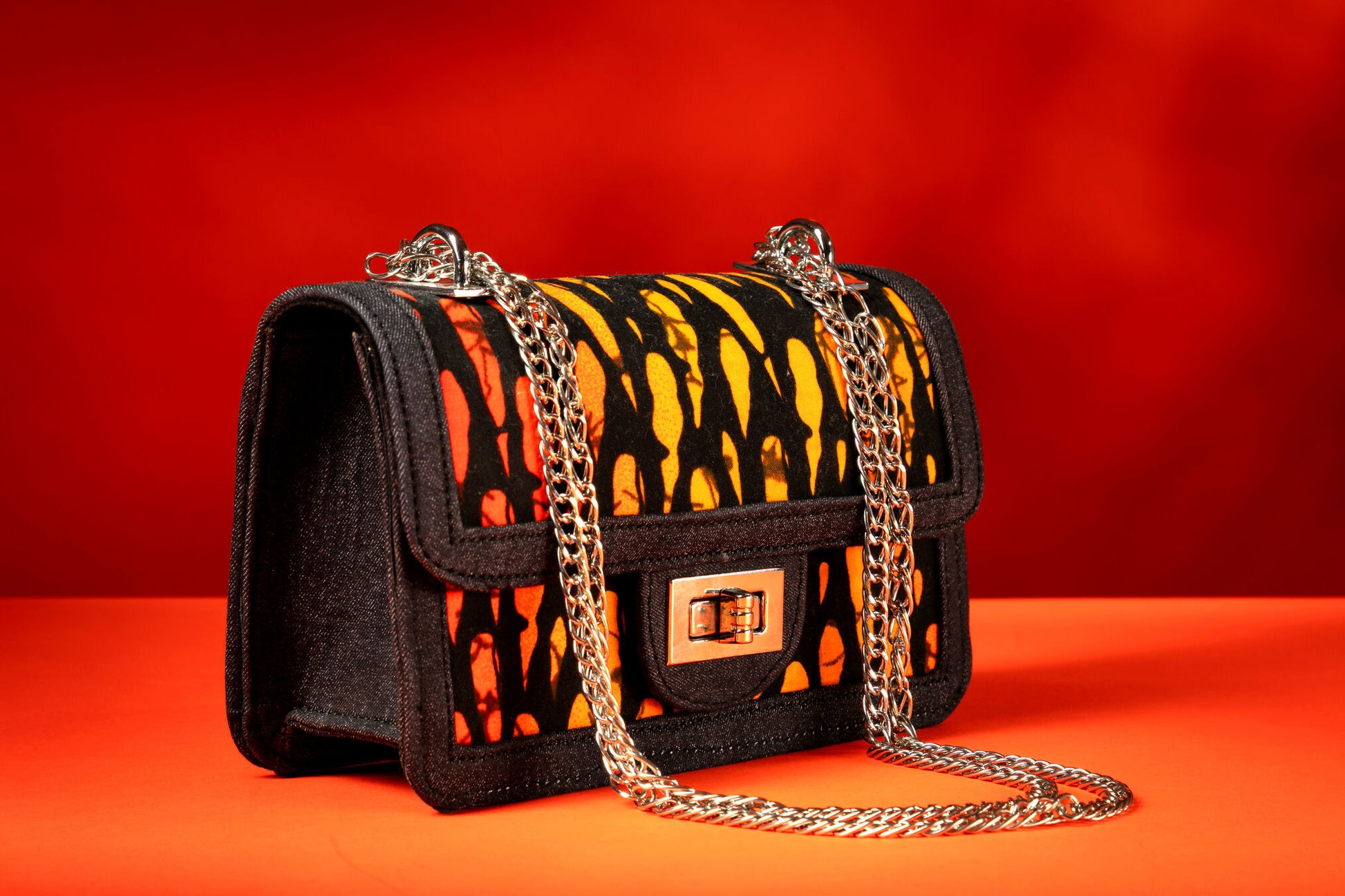 Side angle of Ichaka African print and denim bag with bold orange and black Ankara pattern, accented by a silver chain strap.