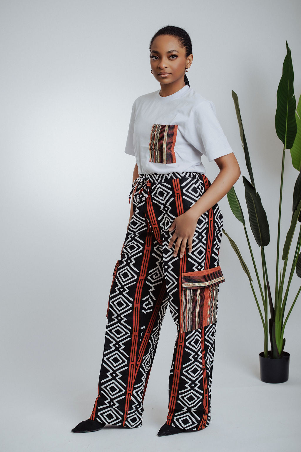 Woman wearing a white t-shirt with a patterned pocket and geometric African  print cargo pants standing next to a plant.