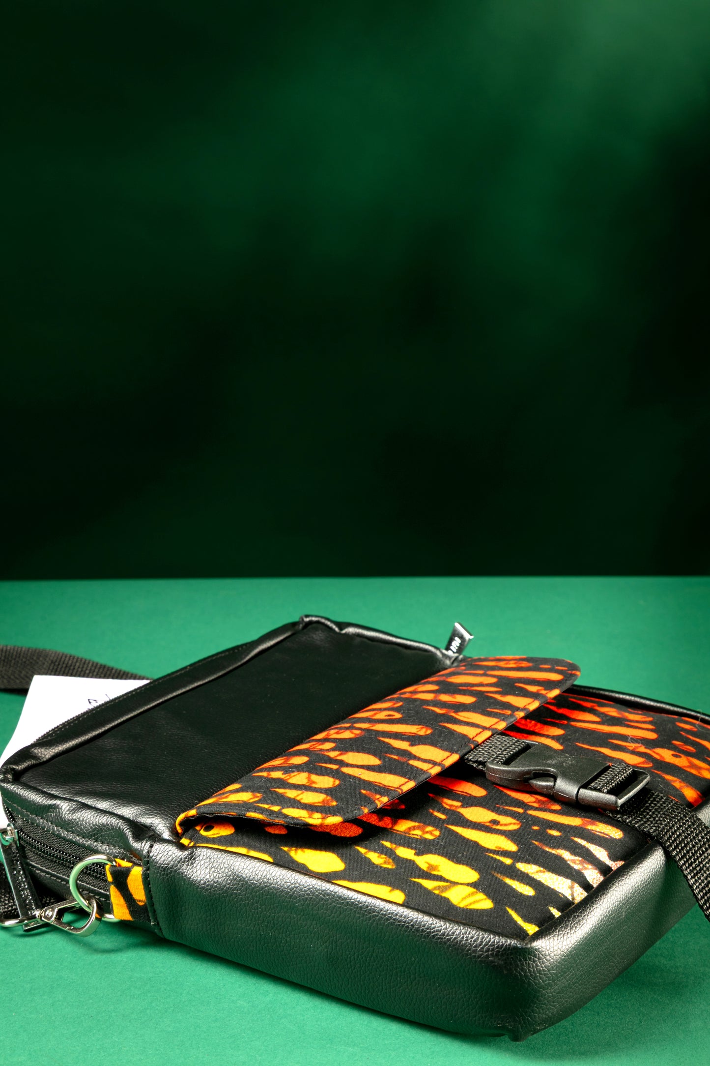 Flat lay of the Krar African Print Crossbody Bag featuring black faux leather and vibrant orange Ankara fabric, with a front flap pocket and buckle strap