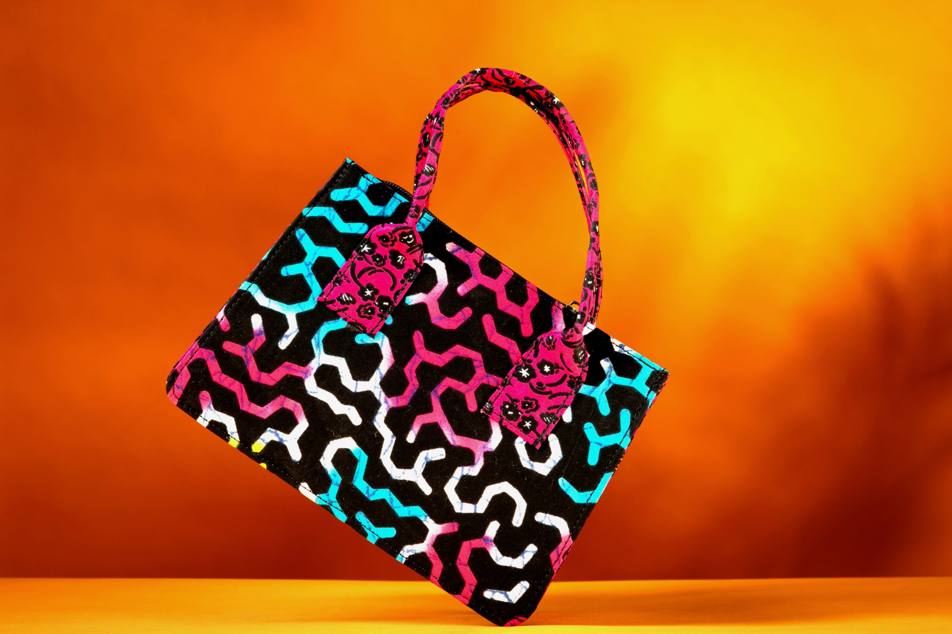 Front view of the Ncegele Bag – a structured African print handbag featuring a multicolor design with contrasting pink patterned handles, photographed against a warm orange background.