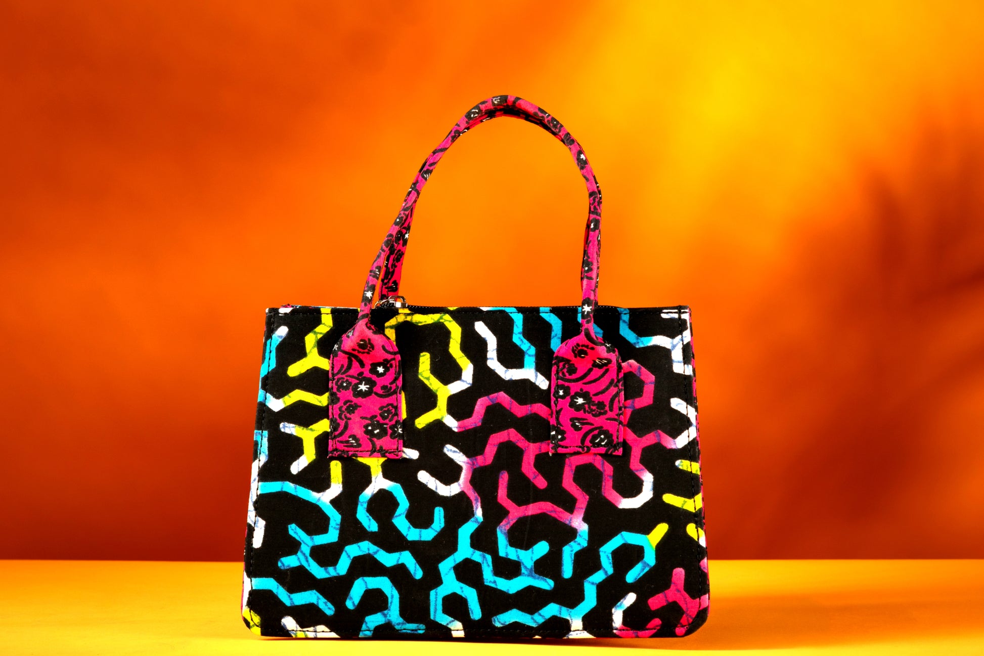 Front view of the Ncegele Bag – a structured African print handbag featuring a multicolor design with contrasting pink patterned handles, photographed against a warm orange background.