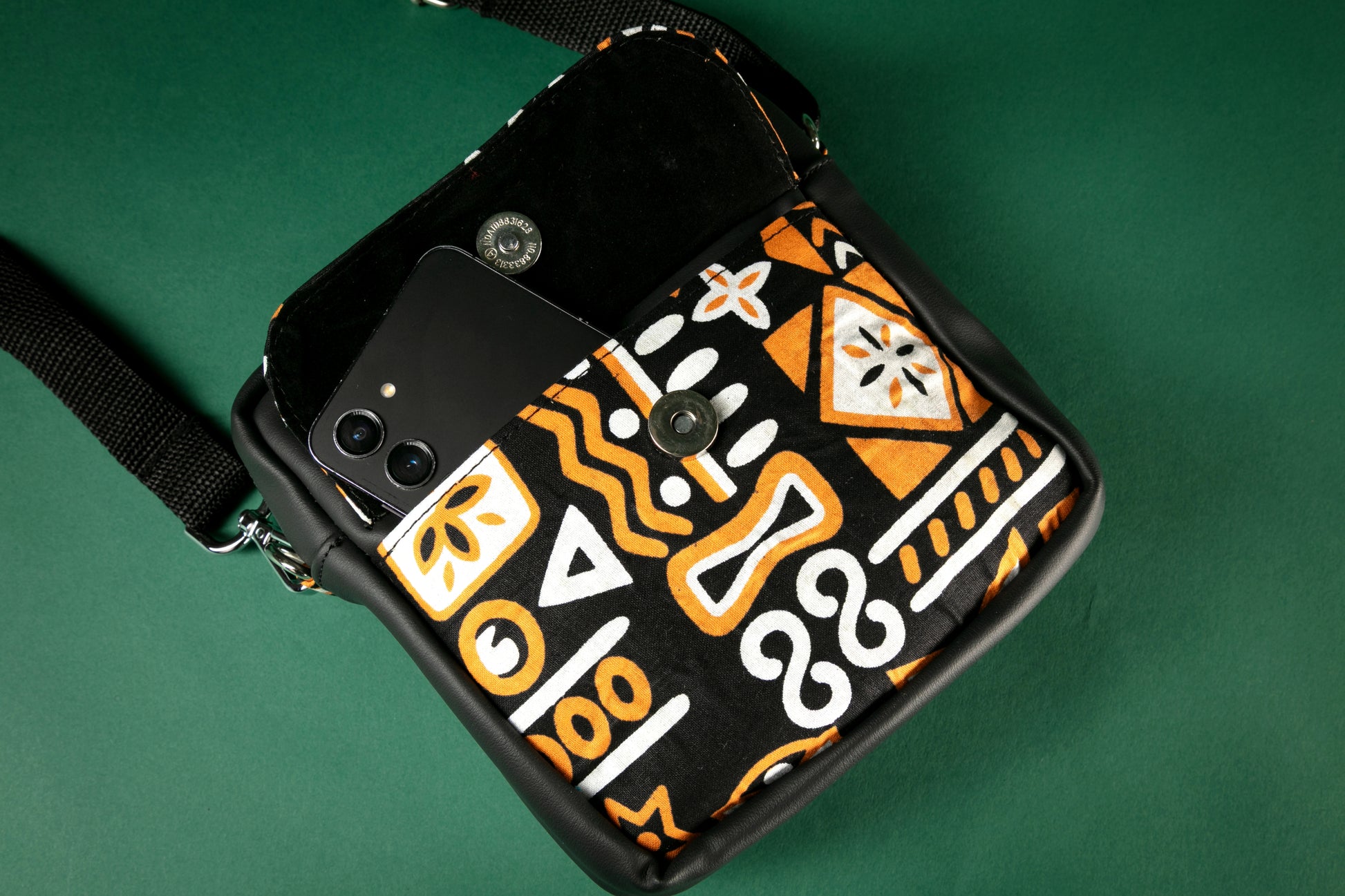 Flat lay of a black crossbody bag with brown, black & white tribal patterns with the flap to the front pocket opened on a green background