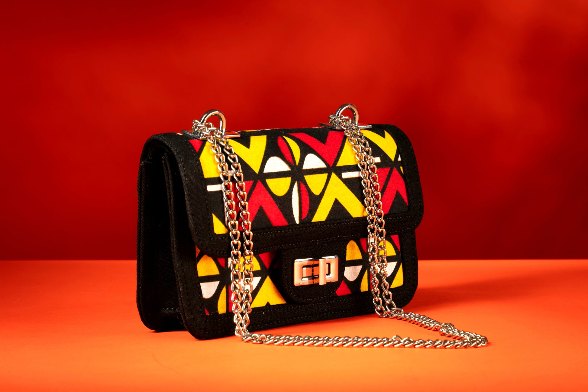 Side angle of Shekere African print and denim bag with bold red, yellow, white, and black Ankara pattern, accented by a silver chain strap.