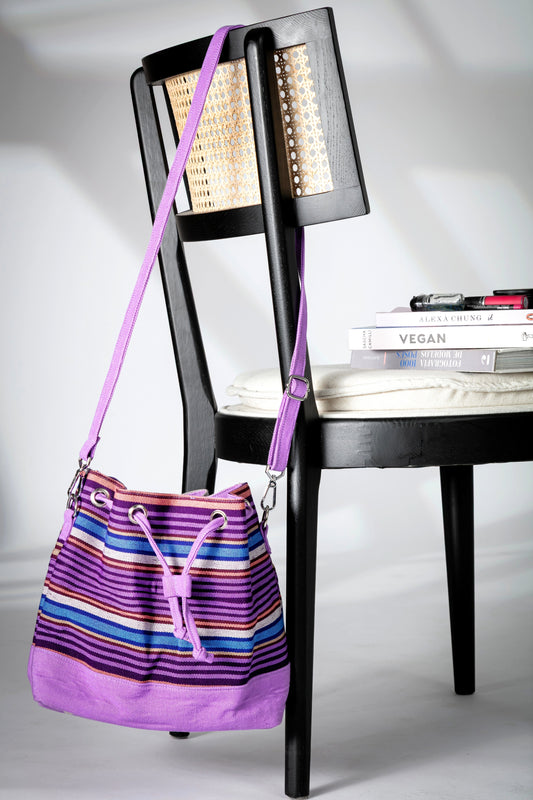 Purple and multicolor striped bucket bag with a long strap and drawstring closure hanging on a chair